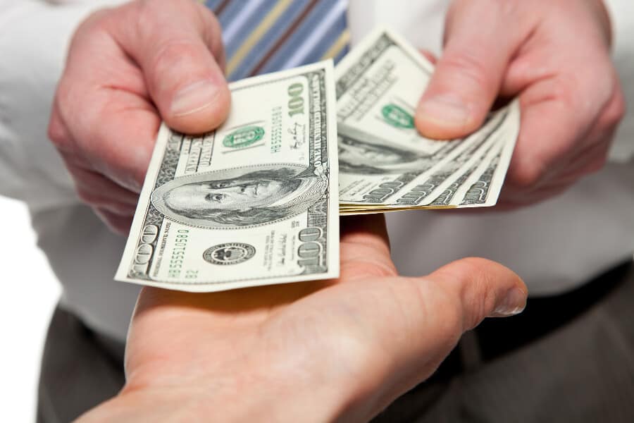 Close up of a man in a tie handing $100 bills to an outstretched hand coming from the camera.