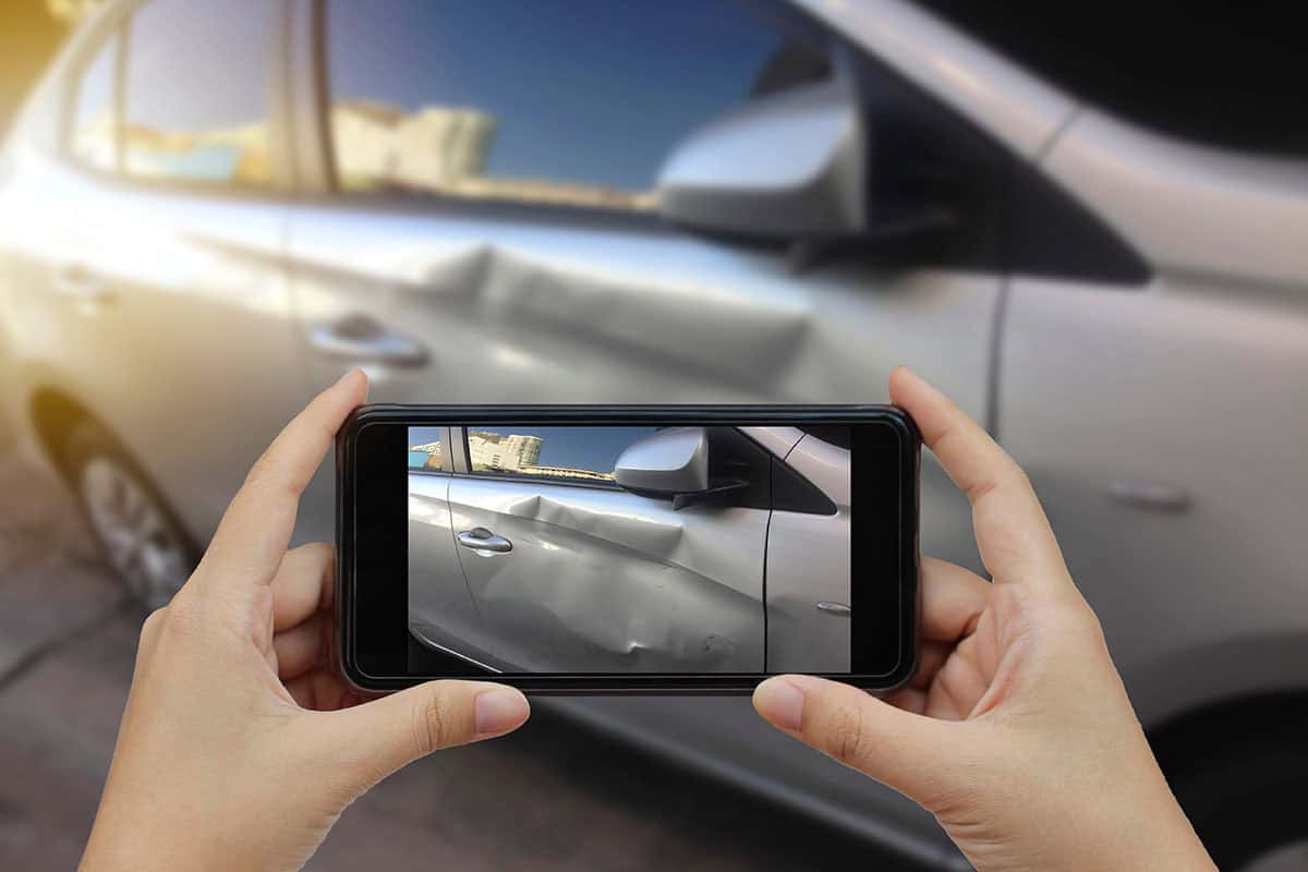 Two hands are holding a phone to photograph a dent on a car door.