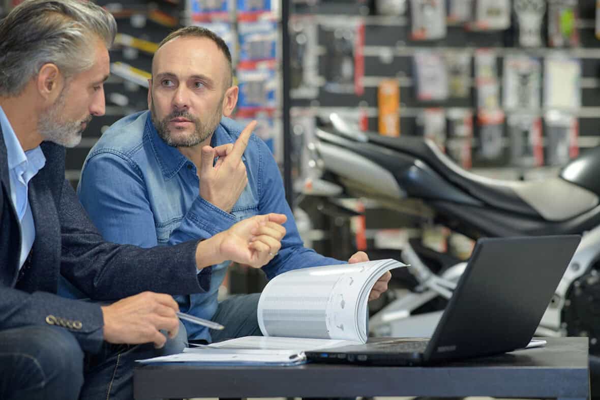 Two men are engaged in a discussion at a table in a motorcycle shop. One man, with grey hair and a beard, listens intently, while the other, with short hair and a beard, gestures while holding a document. A laptop and some paperwork are on the table, and motorcycle parts are visible in the background.