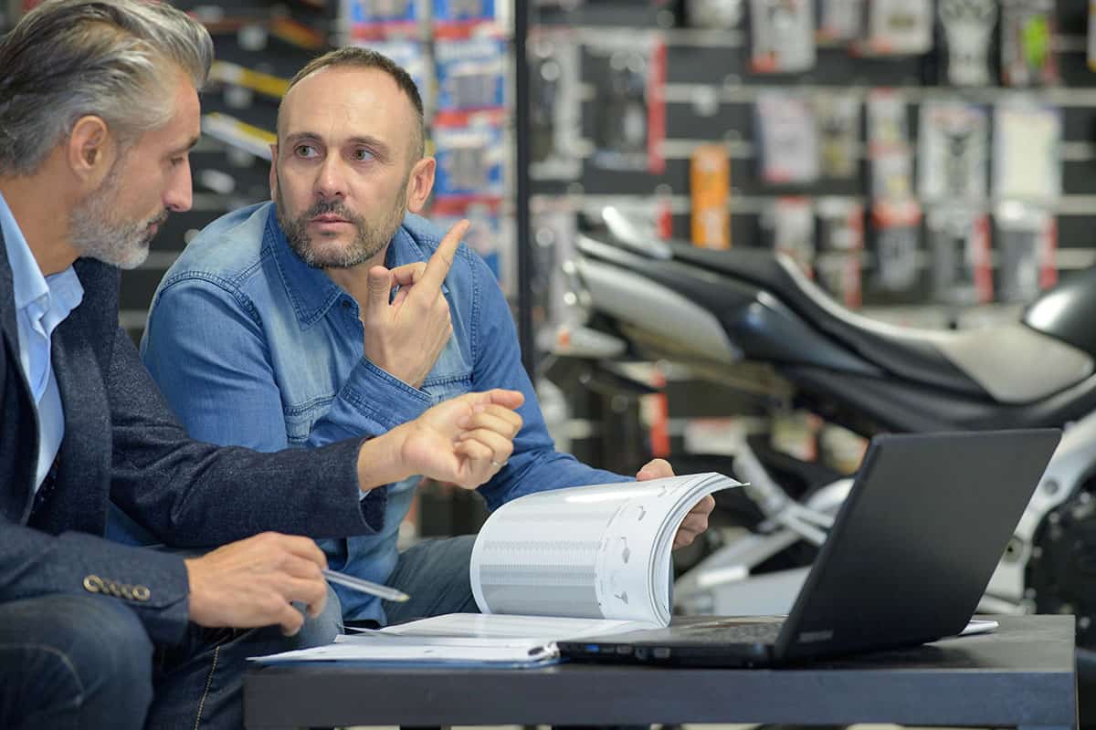 Two men are engaged in a discussion at a table in a motorcycle shop. One man, with grey hair and a beard, listens intently, while the other, with short hair and a beard, gestures while holding a document. A laptop and some paperwork are on the table, and motorcycle parts are visible in the background.