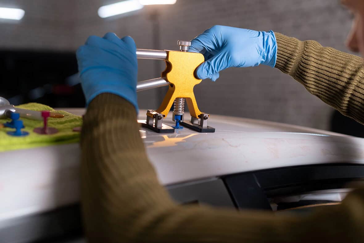 A person wearing blue gloves uses a yellow car dent repair tool on the roof of a car, with colorful suction cups and a green cloth visible nearby.