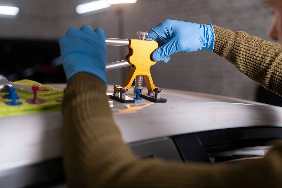 A person wearing blue gloves uses a yellow car dent repair tool on the roof of a car, with colorful suction cups and a green cloth visible nearby.