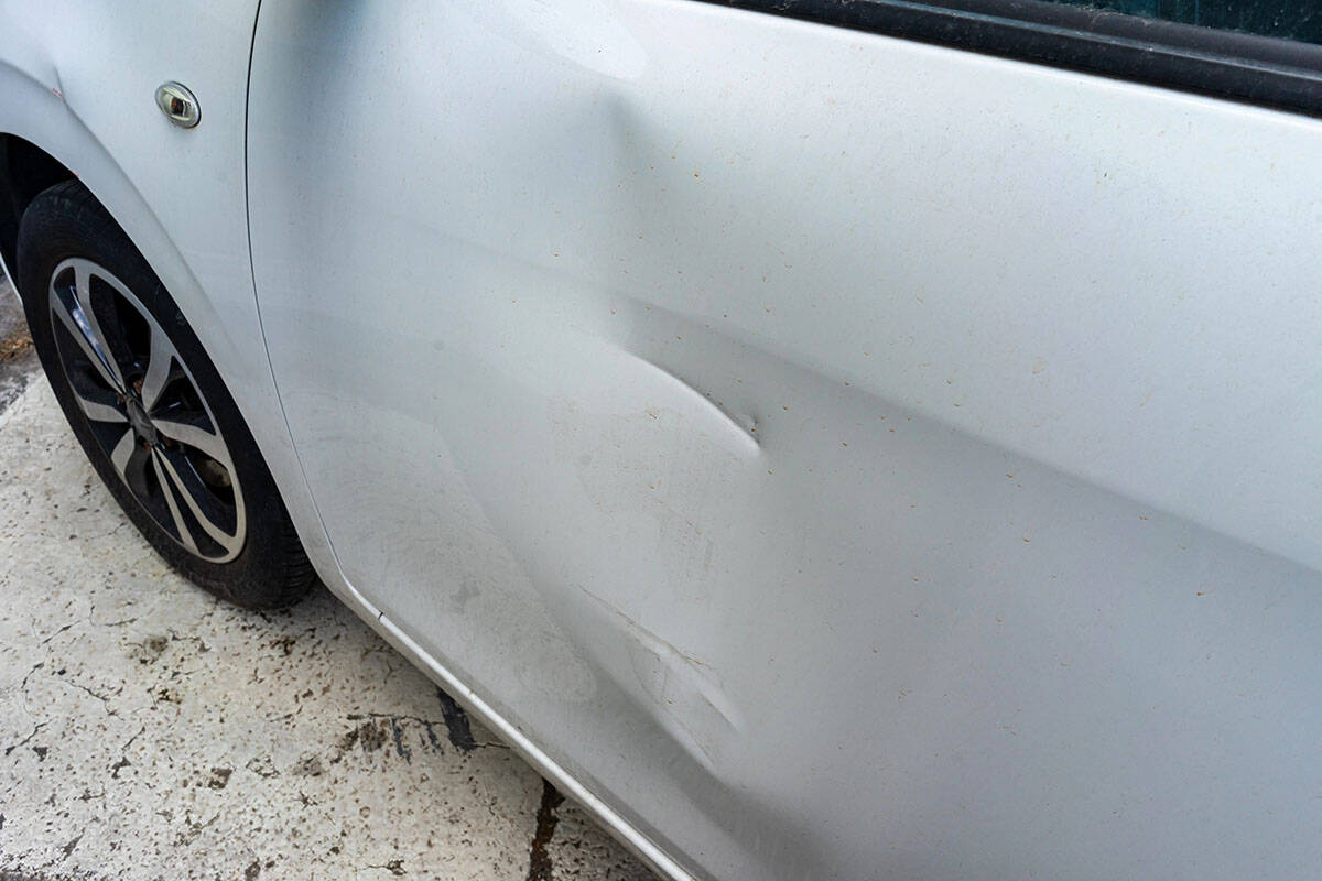 A close-up view of a dent on the side of a white car, with visible dirt and scratches on the surface. The car's wheel is partially visible in the bottom left corner.