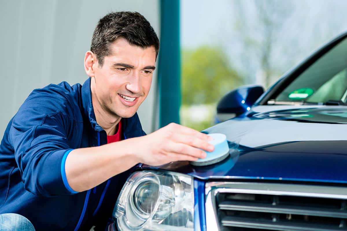 A man in a blue jacket is polishing the hood of a blue car with a circular sponge pad, smiling as he works in a bright, well-lit environment.