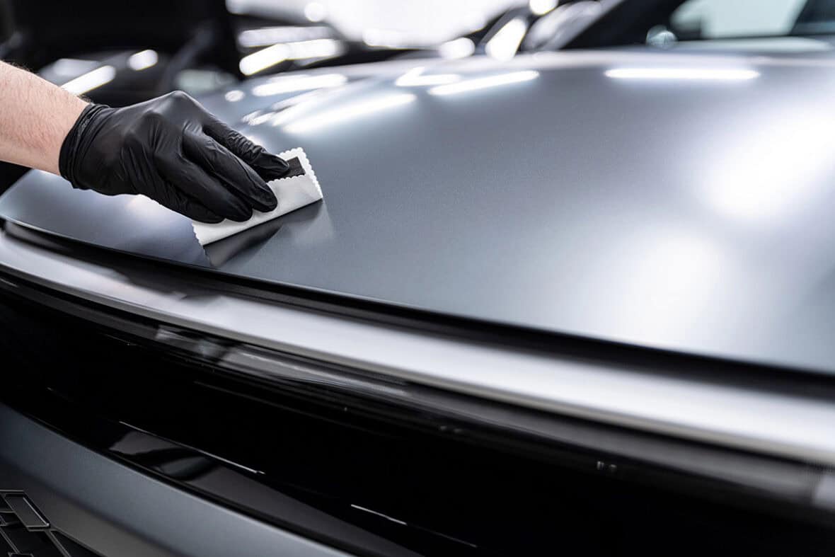 A person wearing black gloves is cleaning the hood of a car with a microfiber cloth, under bright lights in a garage setting.